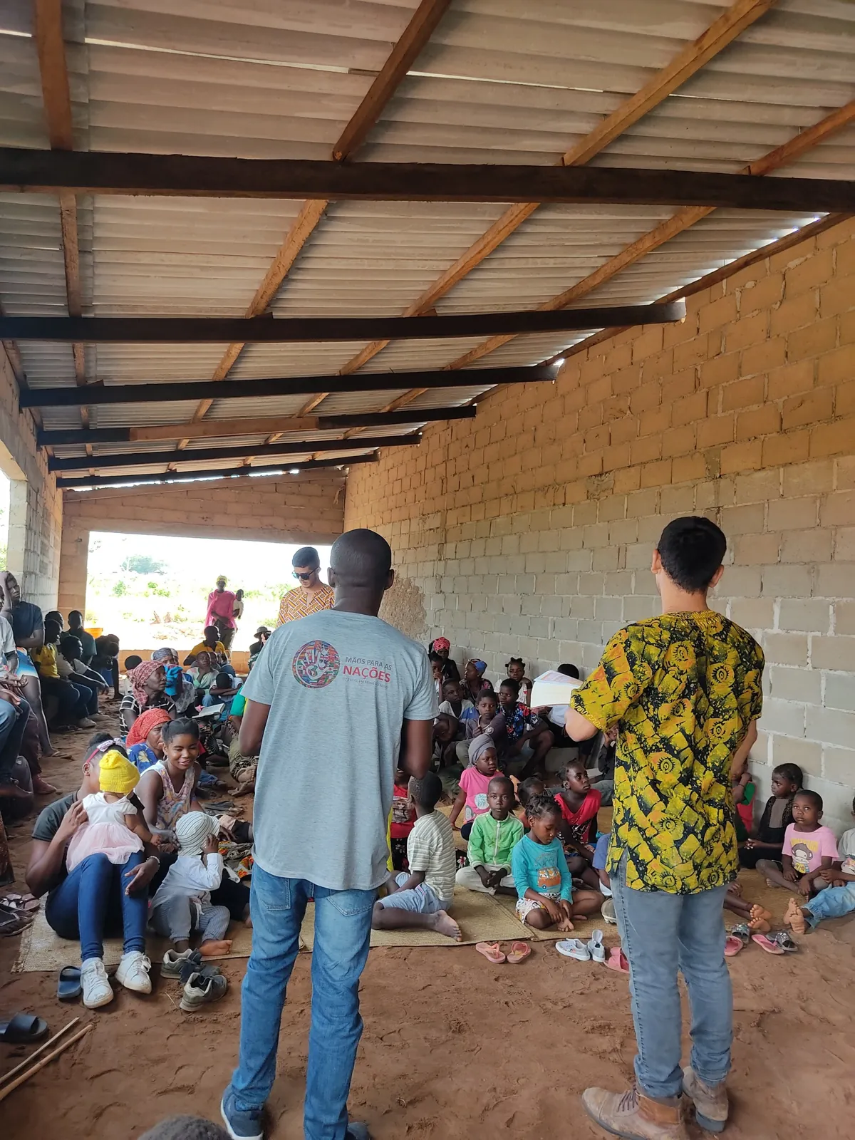 Missionários falando a um grupo grande de crianças e famílias sentadas no chão de um galpão
